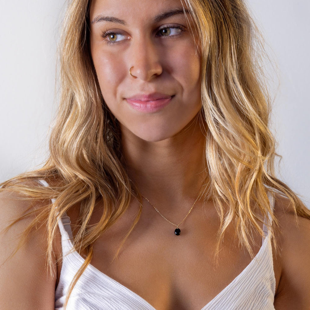May include: A gold necklace with a small, black, oval-shaped pendant. The necklace is delicate and sits close to the neckline. The woman is wearing a white tank top. The woman has long, wavy blonde hair and is looking to the side.